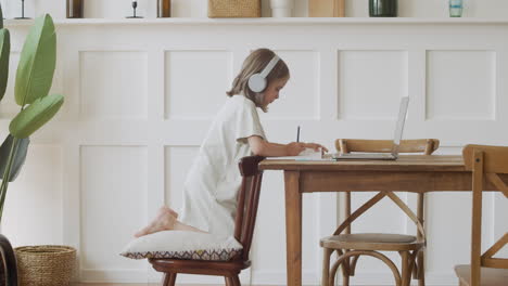 A-Responsible-Cute-Blonde-Girl-With-Headphones-Does-Her-Homework-Writing-On-Paper-While-Looking-At-A-Laptop-Screen