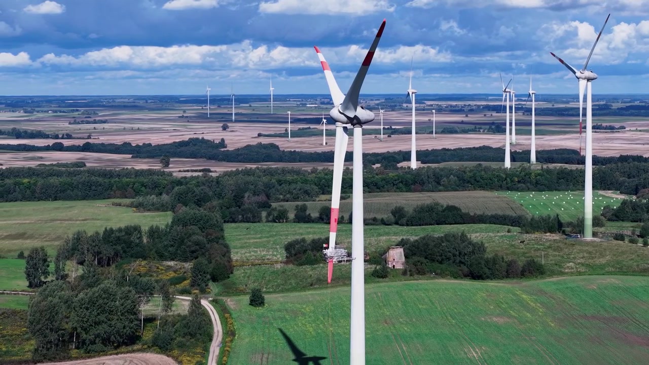 Droneopptak viser moderne vindturbiner som roterer over frodige grønne jorder og jordbruksland under en lys himmel, noe som fremhever fornybar energi i et landlig landskap