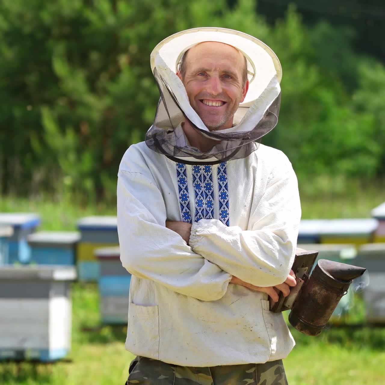 Beekeeper on apiary. Portrait of smiling man on outdoor hives background. Farmer apiarist in protective hat and in embroidery shirt looking on camera in summer day.
