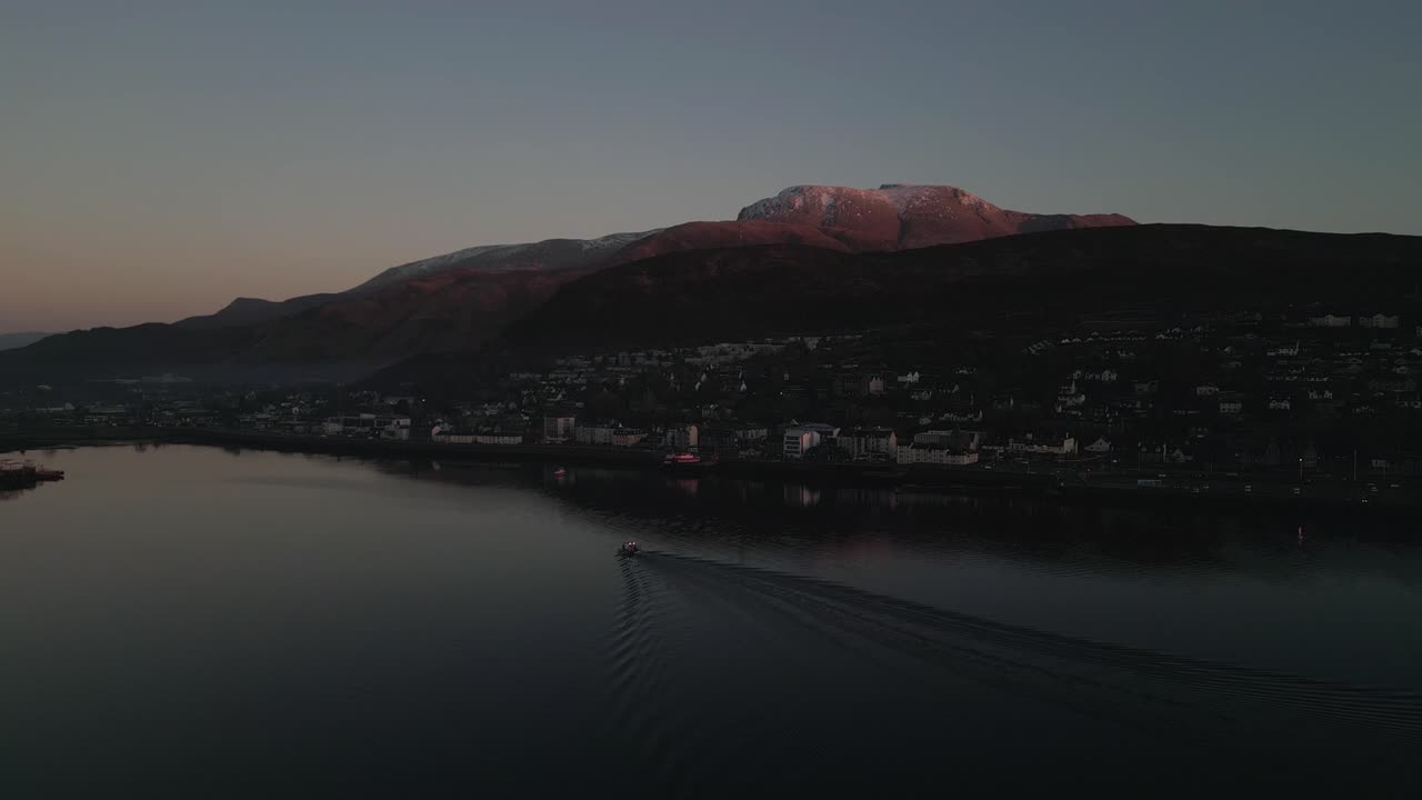 Statisk luftoptagelse af båd, der er på vej til havnen i Fort William med Ben Nevis i baggrunden under solnedgang