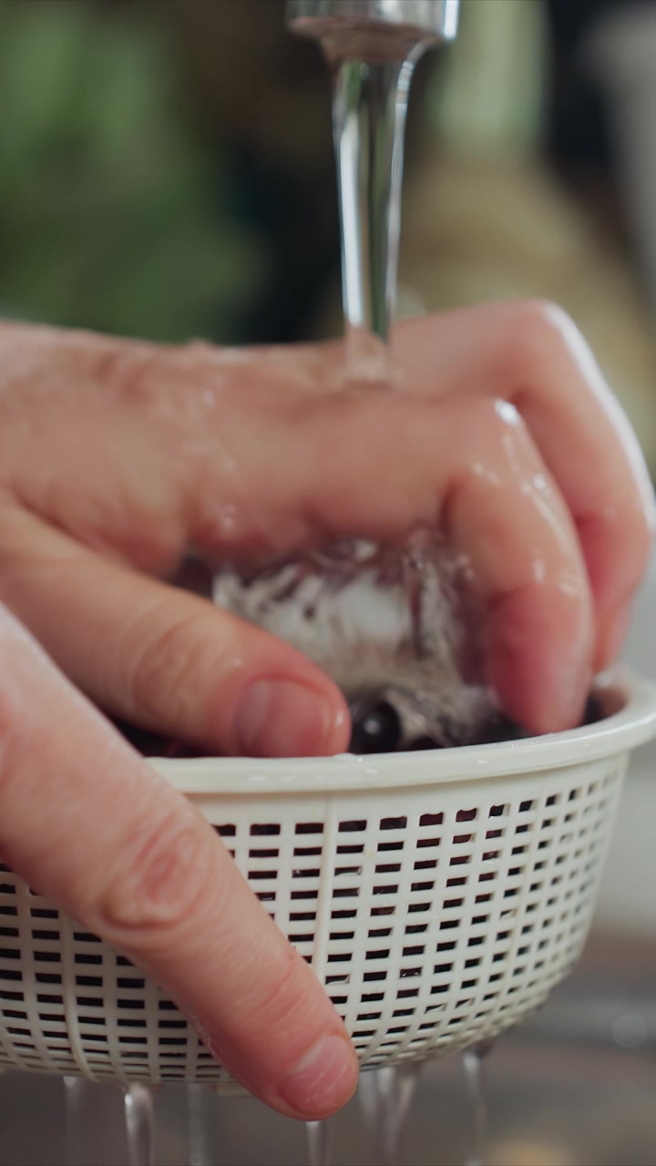 Close-up tangan mencuci ceri gelap di bawah air mengalir. Tangan menggerakkan ceri dan sesekali mengambilnya. Latar belakang termasuk wastafel logam dan meja dapur putih.