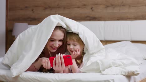 mother lying on bed under duvet with daughter kid girl holding mobile phone watching online movies