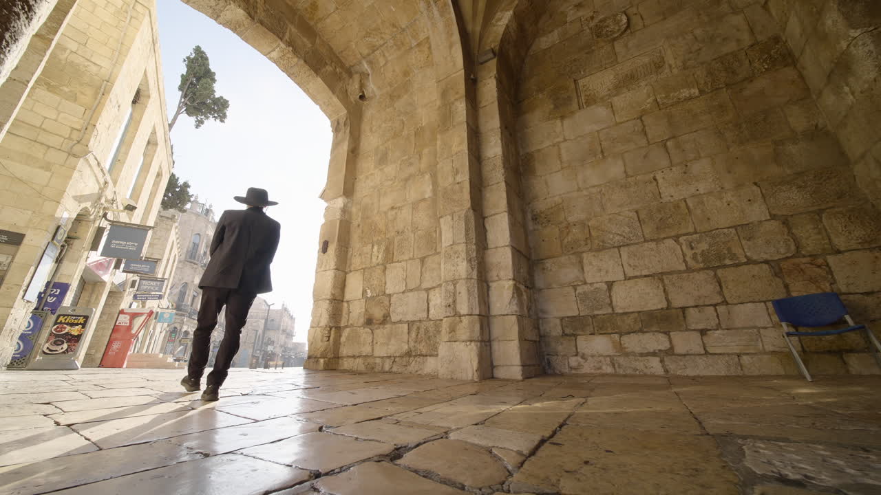 A man in traditional attire walks through a stone archway, stepping into the bright light of the streets of Jerusalem