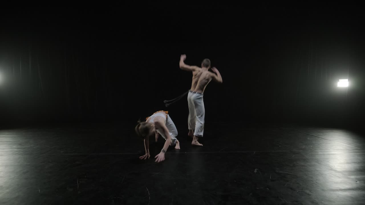 Gruppo di ballerini professionisti che praticano capoeira nell'oscurità contro un riflettore su uno sfondo nero dello studio. Arte marziale afro-brasiliana che combina elementi di danza