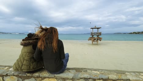 Two-women-in-winter-are-sitting-on-the-wall-at-the-beach