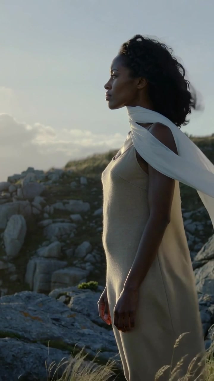 Vertical video: Standing woman in beige dress turning left at horizon as wind lifts scarf, boulders