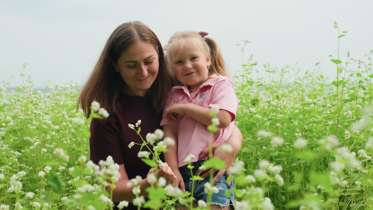 Madre e hija blancas caminan por un prado entre altas flores tomadas de la mano; la madre guía a la pequeña exploradora, cielo suavemente nublado, paisaje rural panorámico, sonrisas amables, cámara lenta natural, afectuoso