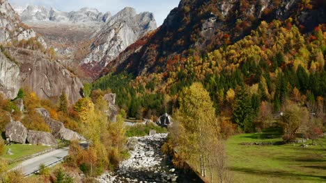 Toma-Aérea-De-Un-Paisaje-Otoñal-Con-árboles-Amarillos,-Verdes-Y-Rojos,-Un-Río-Rocoso-Y-Montañas-En-El-Horizonte,-En-Val-Di-Mello-En-Lombardía,-Italia