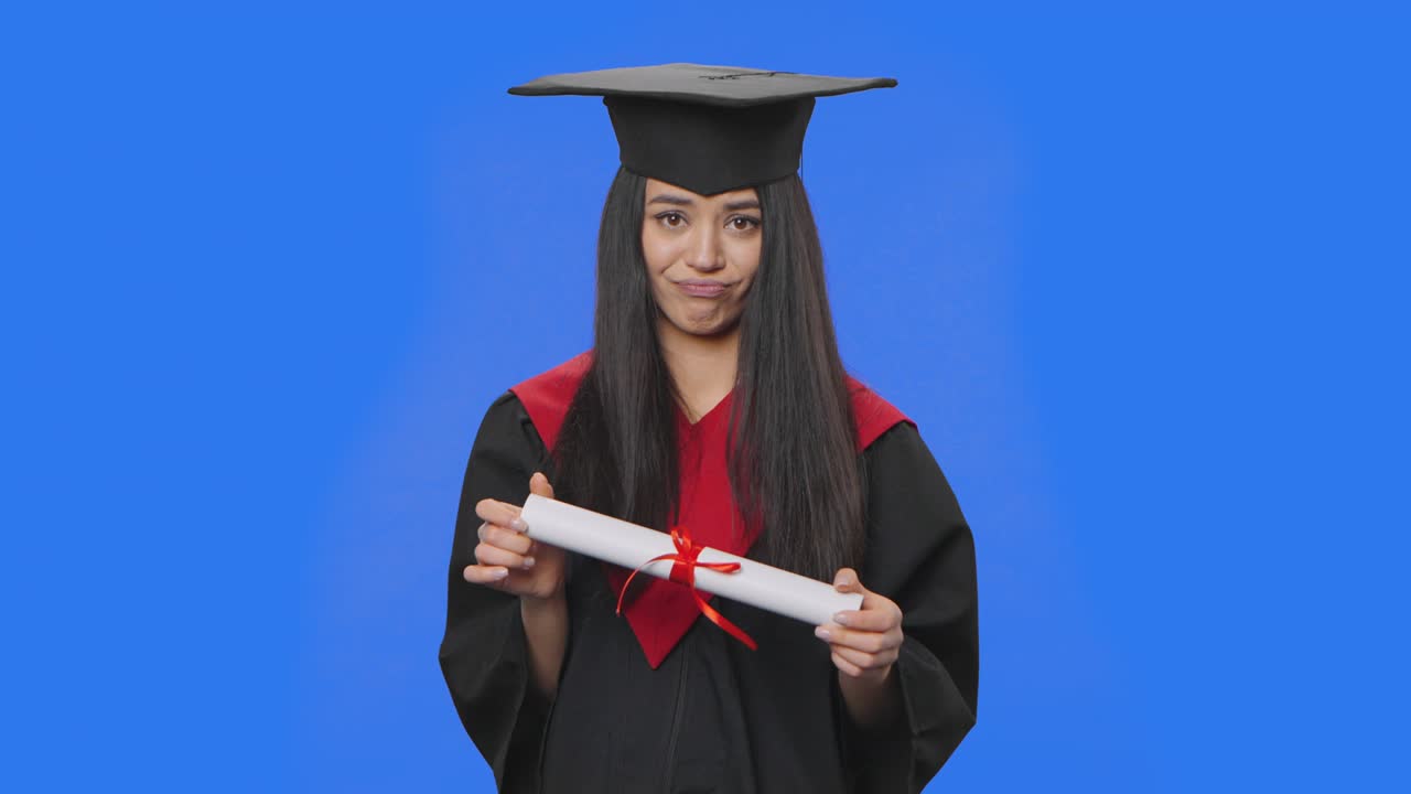 retrato de una estudiante en traje de graduación de gorra y túnica, sosteniendo su diploma y está muy molesta. joven posando en el estudio con fondo de pantalla azul. de cerca. cámara lenta lista 59.94fps