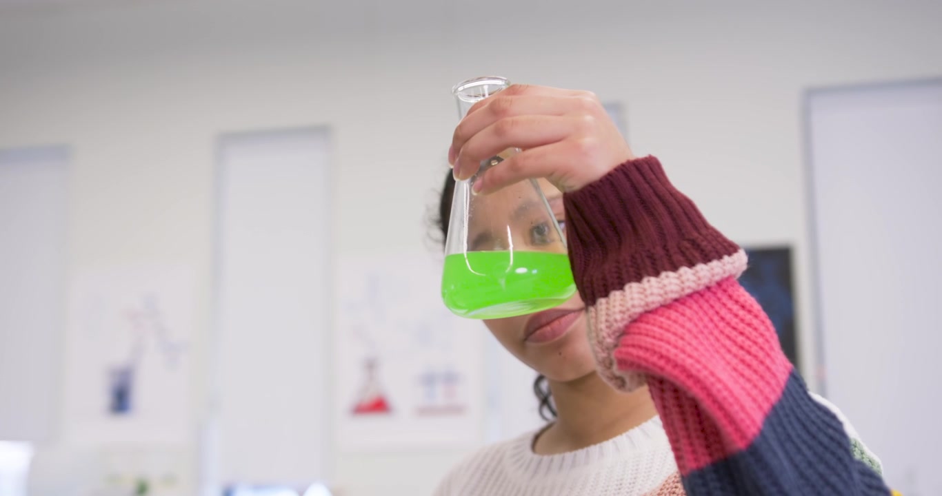 Alunna che, dopo l'indicazione del tablet, solleva e inclina una beuta per ispezionare una soluzione verde nel laboratorio scolastico