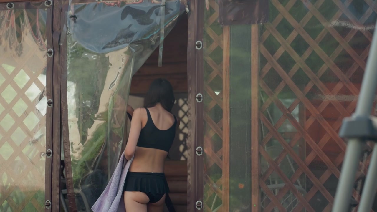 Une femme en brassière de sport noire et short s'approche et ouvre la porte d'une structure en bois extérieure recouverte de bâches en plastique. Elle tient une serviette alors qu'elle entre ou sort de l'espace, révélant des reflets sur sa surface.