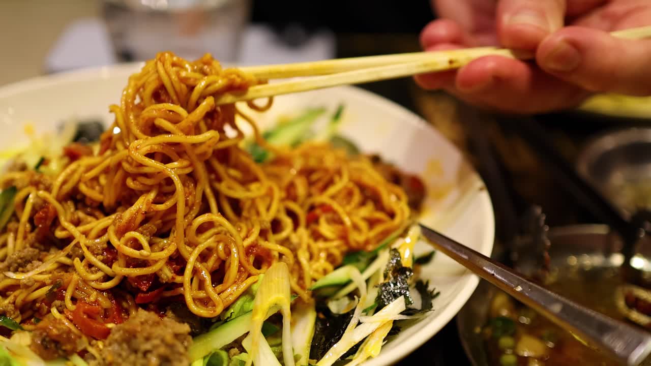 Close-up of chopsticks lifting noodles from a colorful plate with vegetables and garnish.