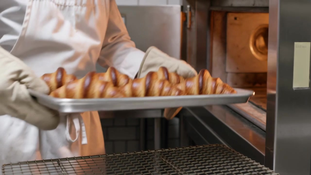 Otevírání trouby, pekař v bílém plášti a zástěře s chňapkami vysouvá plech v pekárně a kontroluje croissanty.