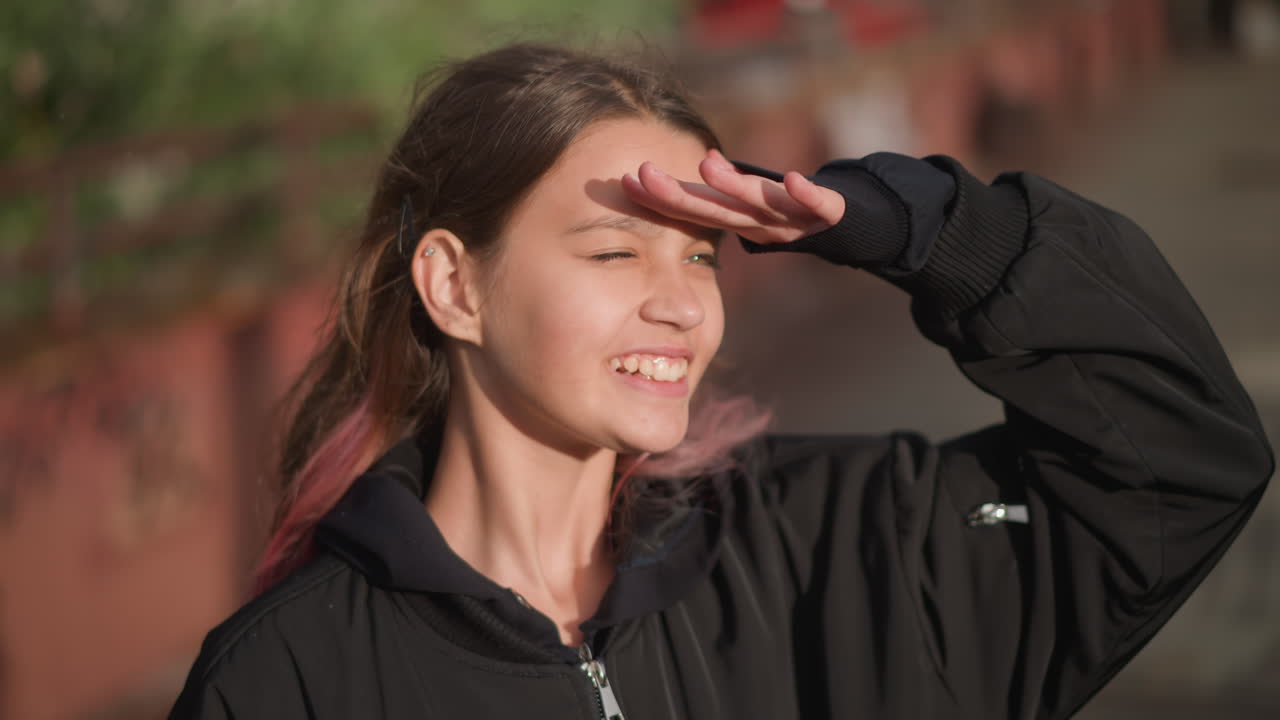 Chica protegiéndose los ojos del sol. Joven con cabello rosa se relaja en un puente lluvioso bajo el sol. Mujer con cabello de puntas rosas y chaqueta informal disfruta del sol en un puente húmedo con sombras largas.
