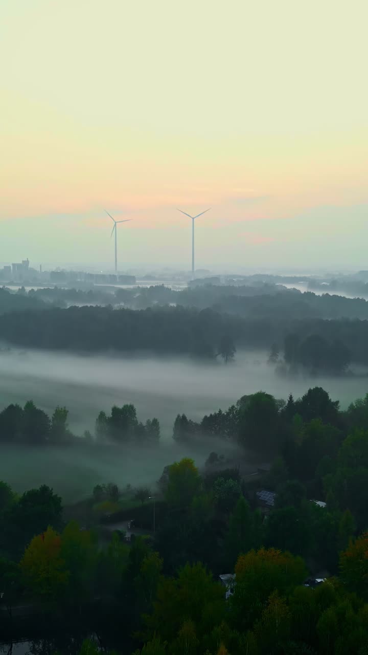 Slow vertical aerial shot flying forward over a rural valley covered in morning fog with wind turbines on the horizon at dawn