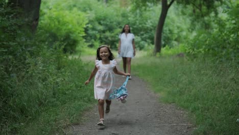 a pregnant woman and her young daughter, both dressed in white, walk hand-in-hand through a grassy park. the scene captures a serene moment of family bonding in nature, surrounded by trees.