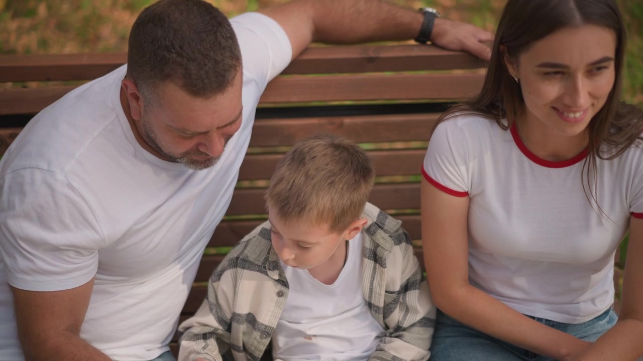 Primo piano di una famiglia seduta insieme su una panchina di legno all'aperto. Un giovane ragazzo tiene un libro aperto mentre i suoi genitori lo guardano, creando una scena calda e coinvolgente con luce naturale sullo sfondo.