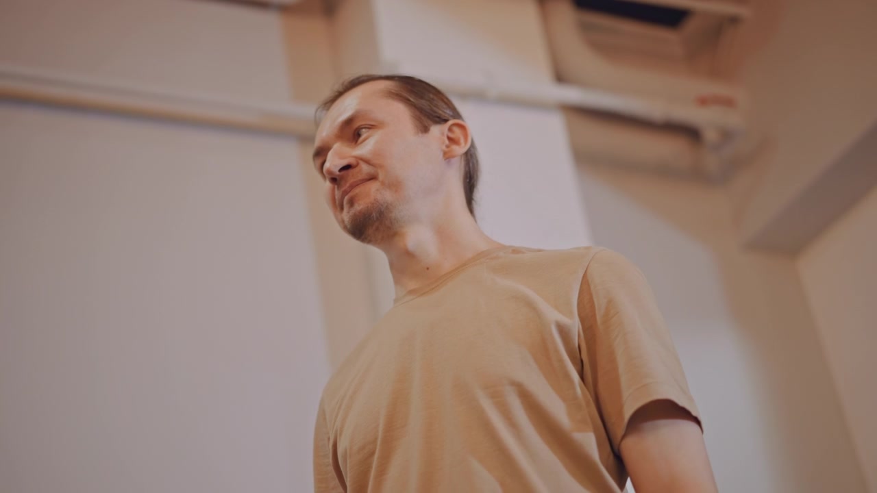Profesor de música (hombre) en estudio dirigiendo un ensayo vocal relajado, de pie bajo tuberías de techo expuestas, con iluminación cálida y suave, camiseta informal, expresión atenta, demostrando técnica de respiración