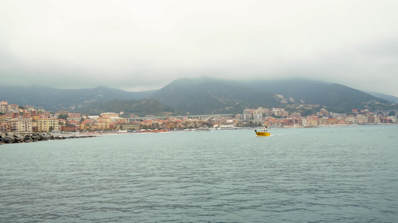Piccola barca gialla naviga nella baia del porto di Varazze in una giornata nuvolosa. Sfondo: città di Varazze, Liguria, Italia