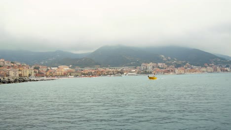 Pequeño-Barco-Amarillo-Navega-En-La-Bahía-Del-Puerto-De-Varazze-En-Un-Día-Nublado,-Ciudad-De-Fondo-Varazze,-Liguria-Italia