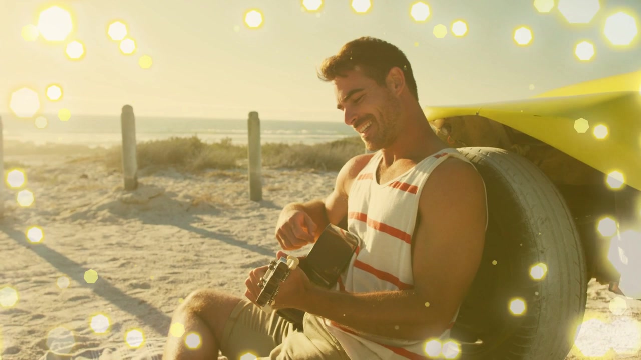 Músico masculino sentado en la playa digitando y luego rasgueando la guitarra, relajándose con efecto bokeh alrededor.