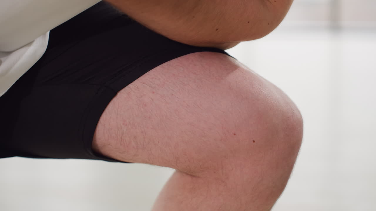 Body built man hands pressing thigh during deep side lunge exercise in bright gym with large windows modern design on light wooden floor wearing white tank top and black shorts focused motion