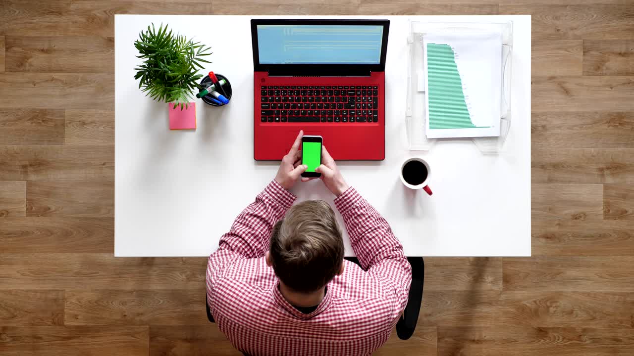 Ung mand taster på telefon med chroma key, topbillede, siddende bag skrivebord med bærbar computer og kaffe