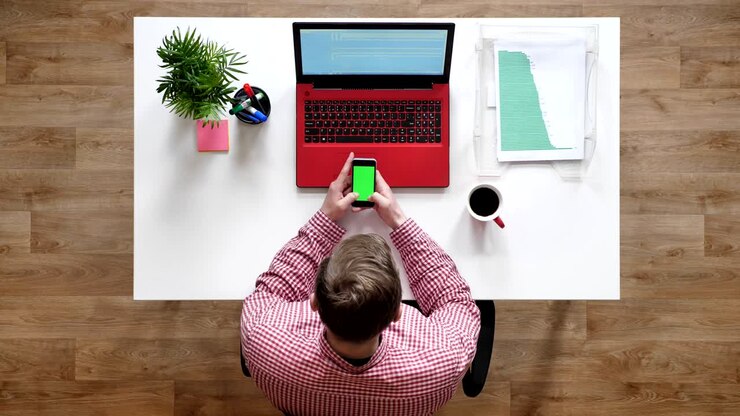 Ung mand taster på telefon med chroma key, topbillede, siddende bag skrivebord med bærbar computer og kaffe
