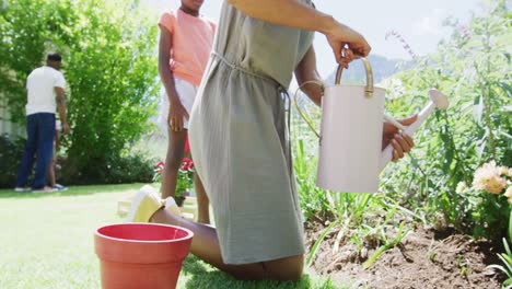 happy african american mother watering flowers in sunny garden, family in background, slow motion