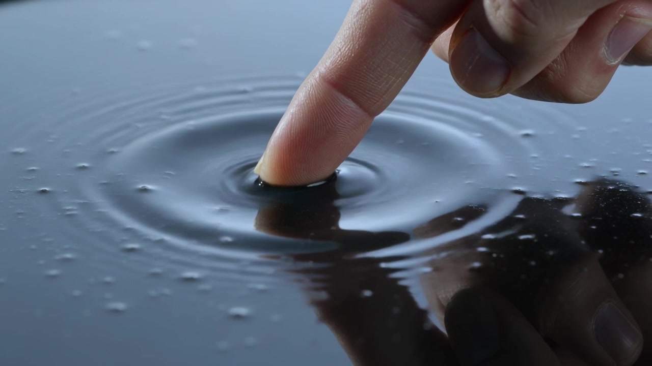 Polpastrello che si avvicina e tocca leggermente una pozza poco profonda, creando increspature circolari e un riflesso.