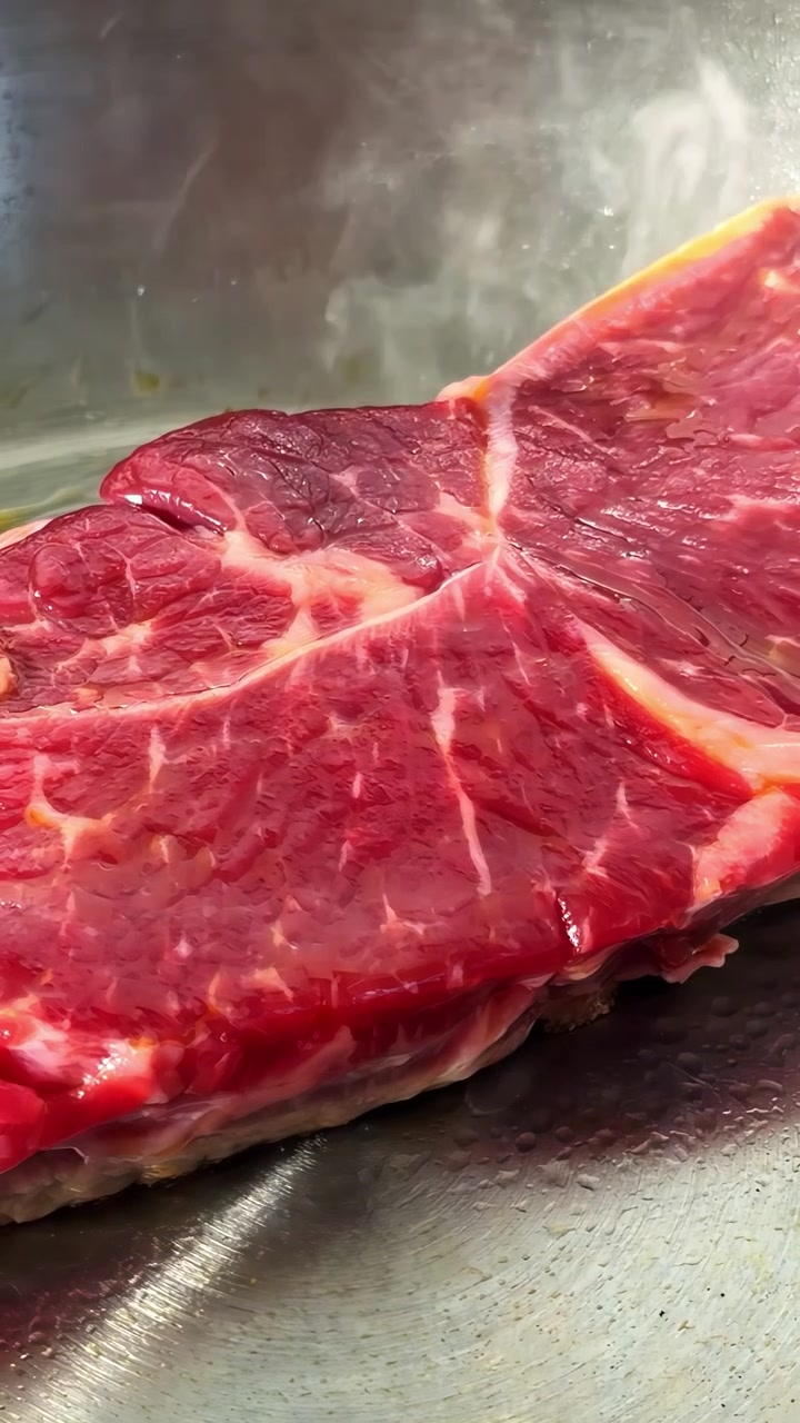 A raw beef steak sizzles gently as it cooks in a hot stainless steel pan, showing fresh marbling and rich red color. Vertical video, ideal for social media.