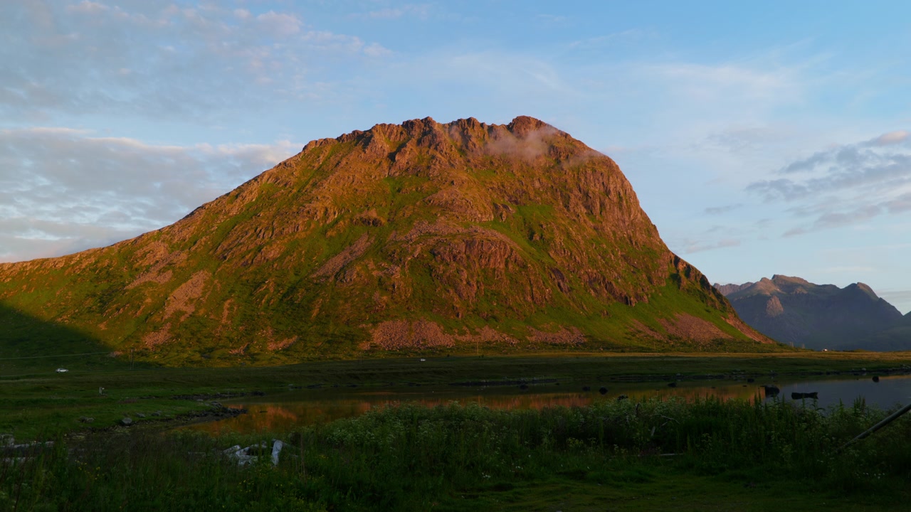 Låg sol lyser upp ett gräsbevuxet berg ovanför en stilla våtmarksdamm i Lofoten, Norge, under en mjuk sommarhimmel