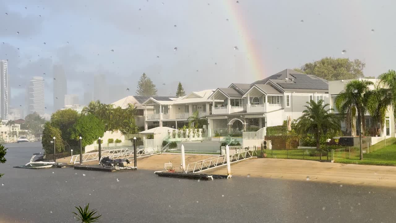 A serene Gold Coast canal scene with luxury homes, a sunshower, and a vibrant rainbow. Soft lighting enhances the tranquil atmosphere