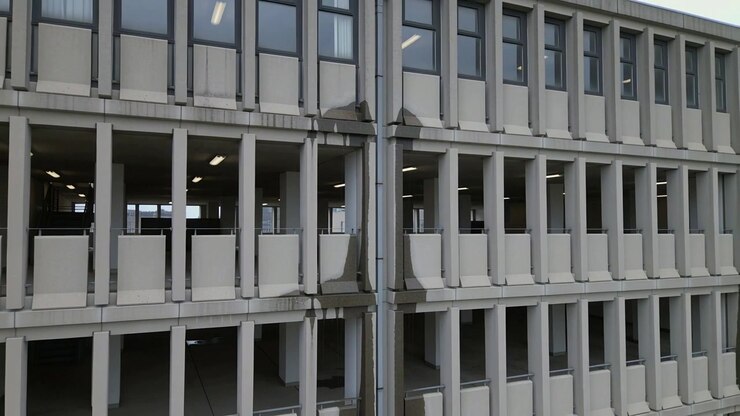La telecamera inizia a panoramare verso destra lungo la facciata del parcheggio per rivelare campate ripetute, pluviali e luci