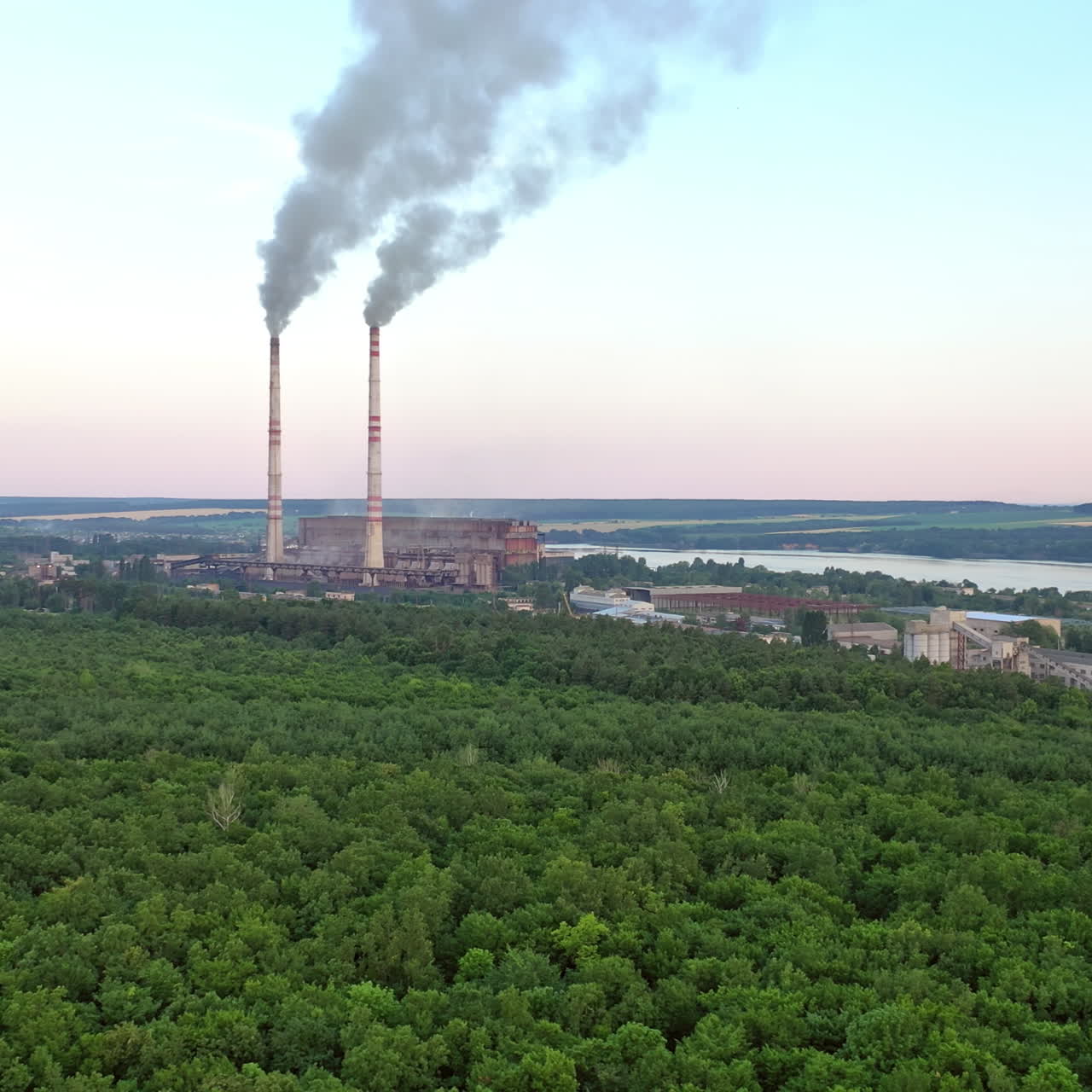 Aerial view on the green forest and pipes with smoke from plant. Natural landscape of woodland near the river and industrial factory on the bank of the river.