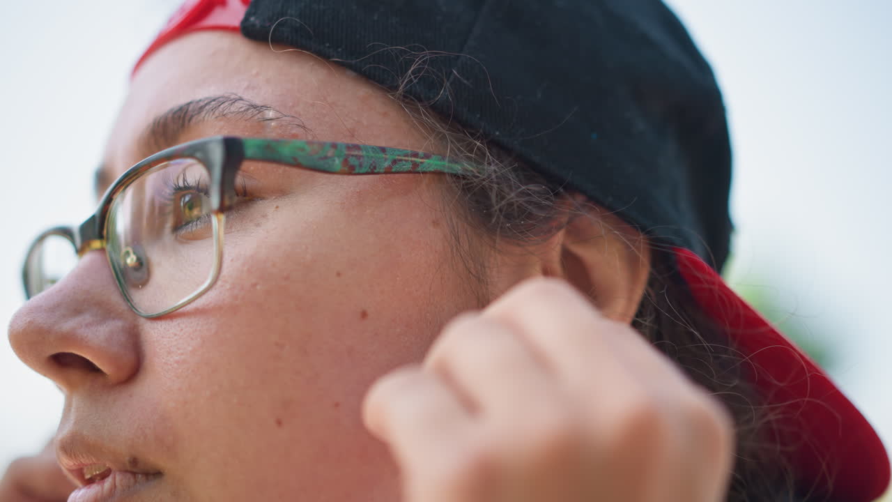 primer plano de una persona disfrutando de la música al aire libre, con la mano tocando el auricular mientras los ojos se enfocan en la distancia, con gafas y gorra visibles, un smartwatch en la muñeca, fondo bokeh suave y expresión serena que transmite audio