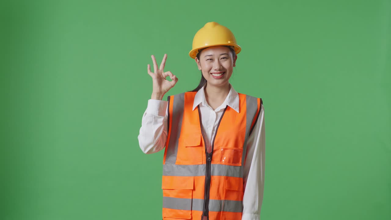 안전 헬을 입은 아시아 여성 엔지니어가 미소 짓고 녹색 스크린 배경 스튜디오에 서 있는 동안 카메라에 은 몸짓을 보여줍니다.