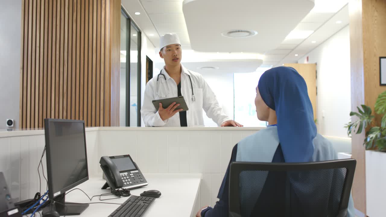 Dokter Asia dengan stetoskop memeriksa jadwal di tablet di meja sementara resepsionis Afrika mendengarkan.