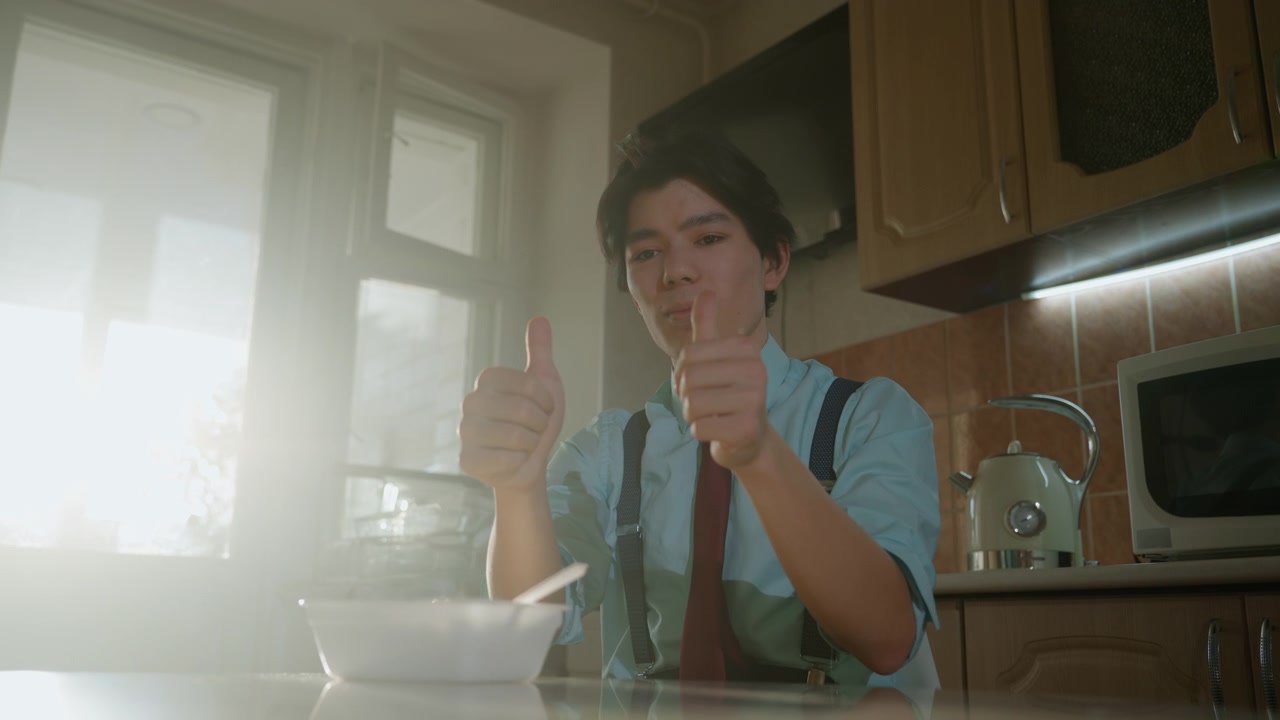 Giovane uomo asiatico che fa colazione in una cucina illuminata dal sole, fa un pollice in su e poi è sorpreso dal sapore, sorriso giocoso e risata di sollievo, che indossa camicia, cravatta e bretelle, un'ampia sequenza mostra la sua espressività