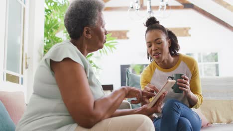 Afroamerikanische-Tochter-Mit-Kaffeetasse-Zeigt-Ihrer-Mutter,-Wie-Sie-Zu-Hause-Ein-Digitales-Tablet-Nutzt