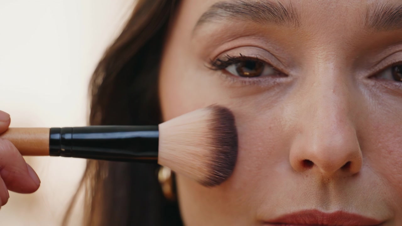 Closeup model applying cosmetic powder with large fluffy brush looking camera.