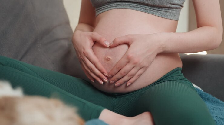Closeup pregnant belly with hands forming heart over bump, green leggings, textured couch foreground, intimate maternal gesture, fetal movement focus, tender prenatal bonding, soft daylight, lifestyle