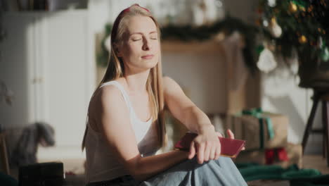 mujer con los ojos cerrados sosteniendo un libro en el sol iluminado en casa durante la navidad