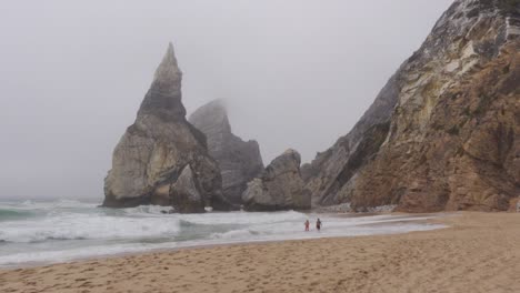 A-Couple-taking-a-dip-at-a-Stormy,-Foggy-Beach-at-Praia-da-Ursa,-Portugal