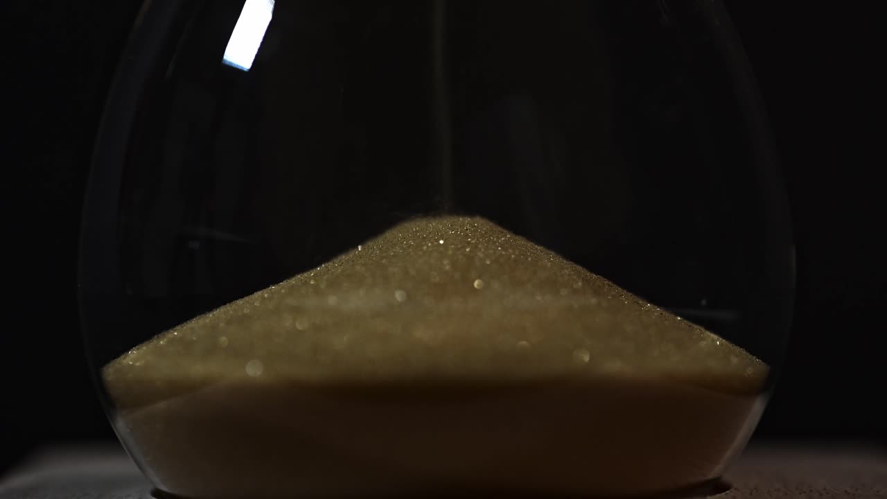 A close-up view of golden sand accumulating in the lower chamber of an hourglass. The dark background highlights the sparkling texture of the fine grains as time slowly passes.