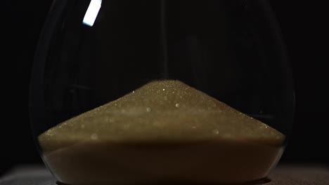 a close-up view of golden sand accumulating in the lower chamber of an hourglass. the dark background highlights the sparkling texture of the fine grains as time slowly passes.