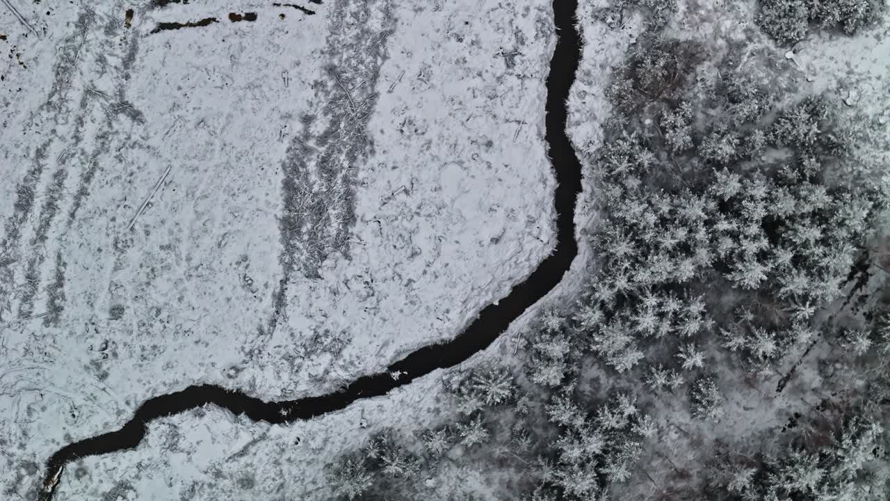 Veduta aerea dall'alto di un torrente tortuoso che si snoda attraverso campi e foreste innevati in inverno. L'acqua scura contrasta con il terreno bianco, formando motivi naturali su un paesaggio rurale ghiacciato