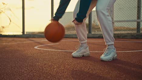 Primer-Plano-De-Una-Niña-Con-Zapatillas-Blancas-Y-Uniforme-Deportivo-Golpea-Una-Pelota-Naranja-Desde-El-Suelo-En-La-Cancha-De-Baloncesto-De-La-Calle-Roja-Por-La-Mañana.