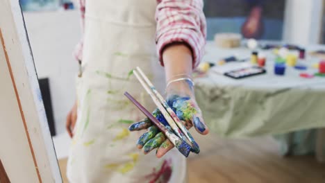 hand of biracial female artist in apron holding paint brushes in art studio, slow motion