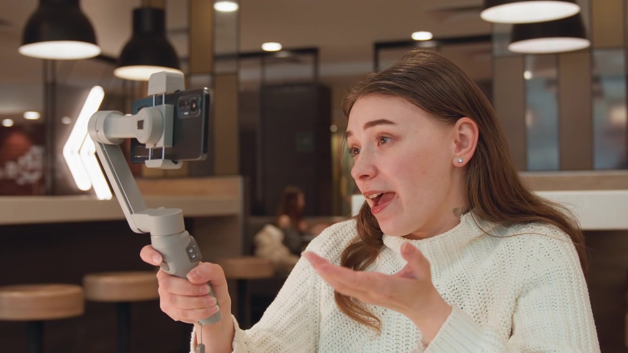 Giovane donna tiene uno smartphone montato su gimbal a braccio teso e gesticola mentre parla con movimenti facciali coinvolti ed espressivi; il movimento mostra lievi rotazioni della testa, tocchi della mano sulla guancia e una presa ferma.